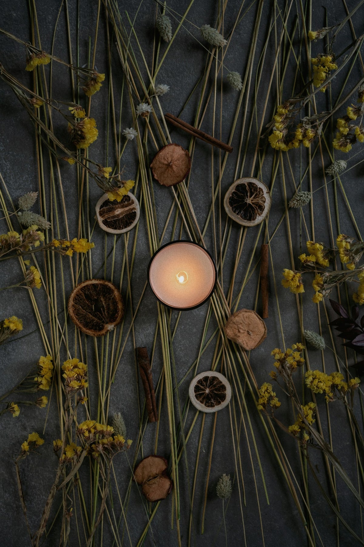 Lit candle with dried flowers, citrus and cinnamon on a dark surface